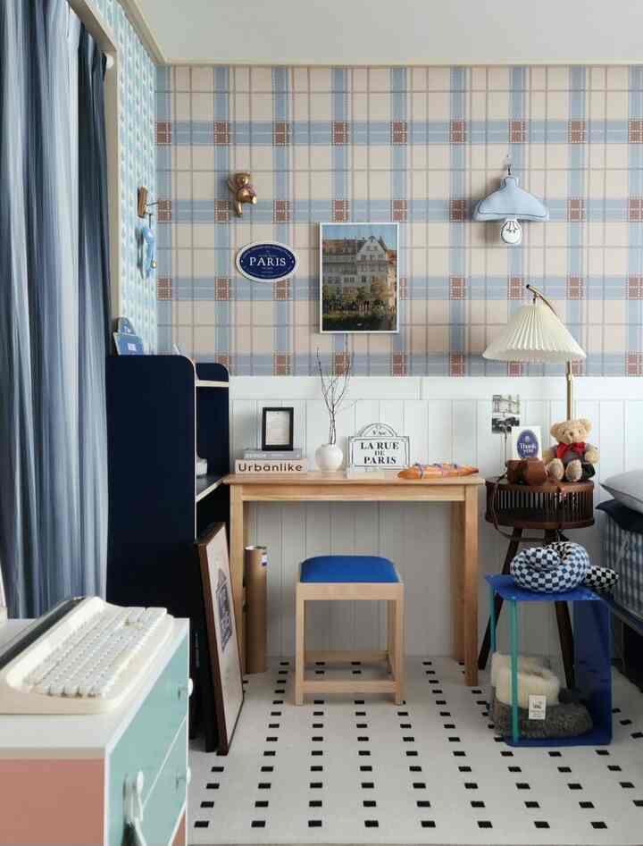 A cozy small room in soft blue tones featuring a wooden desk, blue stool, various wall decorations, and cute stuffed toys