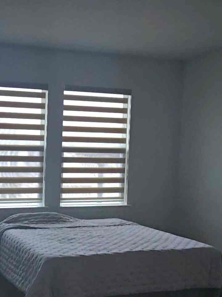 White-toned bedroom featuring a bed with a white quilted duvet cover centrally placed under two striped blinds, creating a clean atmosphere