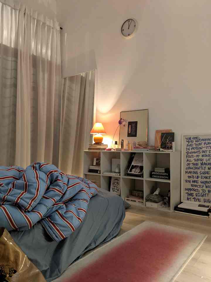 Monotone white and beige small bedroom featuring a bed, bookshelf, and warm orange table lamp creating a cozy atmosphere