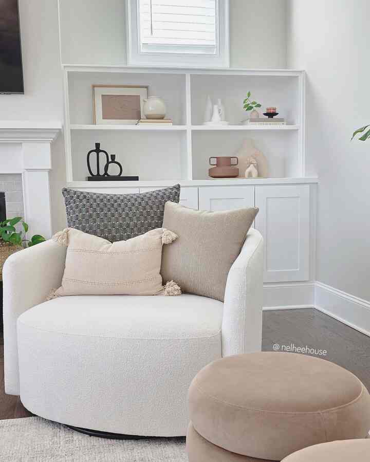 Beige and white toned living room corner featuring a curved armchair, ottoman, cushions, and simple natural decor on wall shelves