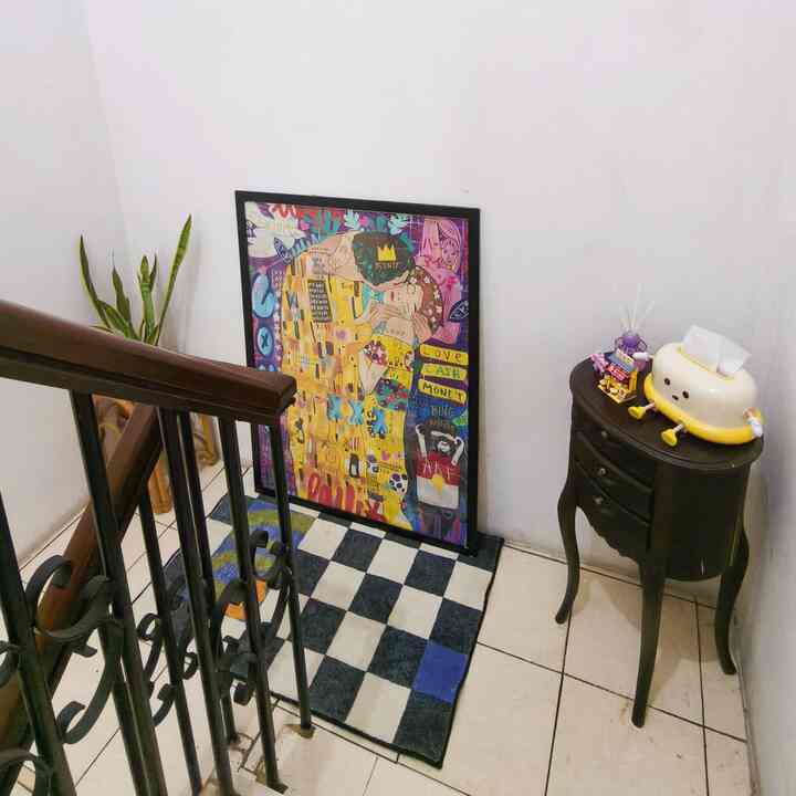 A small stair corner with white walls and black railing featuring a vintage-style curved leg table, checkered mat, and colorful framed artwork, creating a cozy decor spot