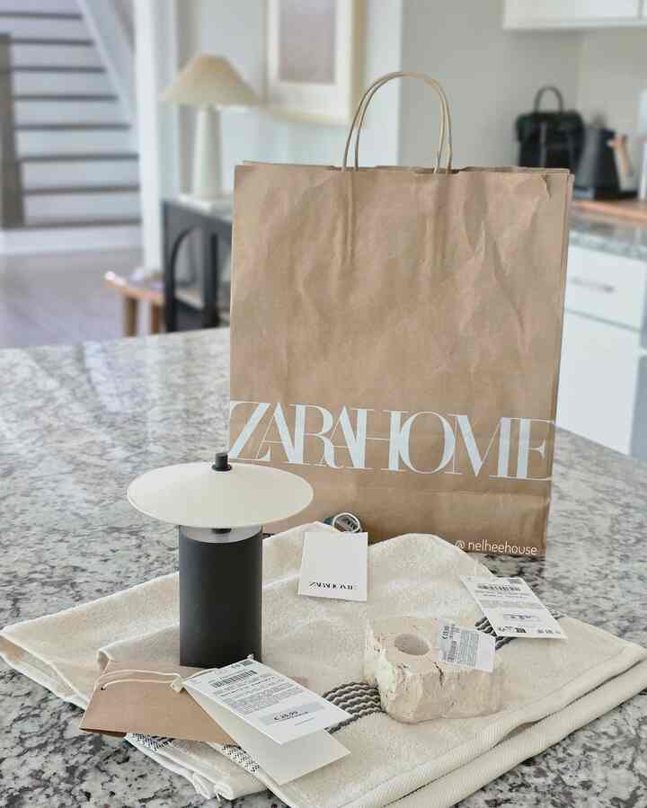A beige and white toned kitchen counter featuring a Zara Home shopping bag, natural material towels, and a candleholder in a simple setting.