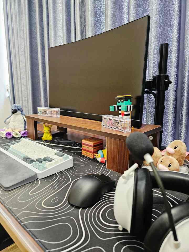 Compact desk with black tones featuring a wooden monitor stand, curved monitor, gaming keyboard, and headset in a home office setup