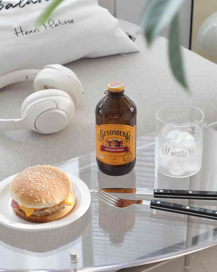 Cafe-style setting with light beige and white tones featuring a cheeseburger on a round plate, Bundaberg ginger beer bottle, glass with ice, and white headphones on a minimal table