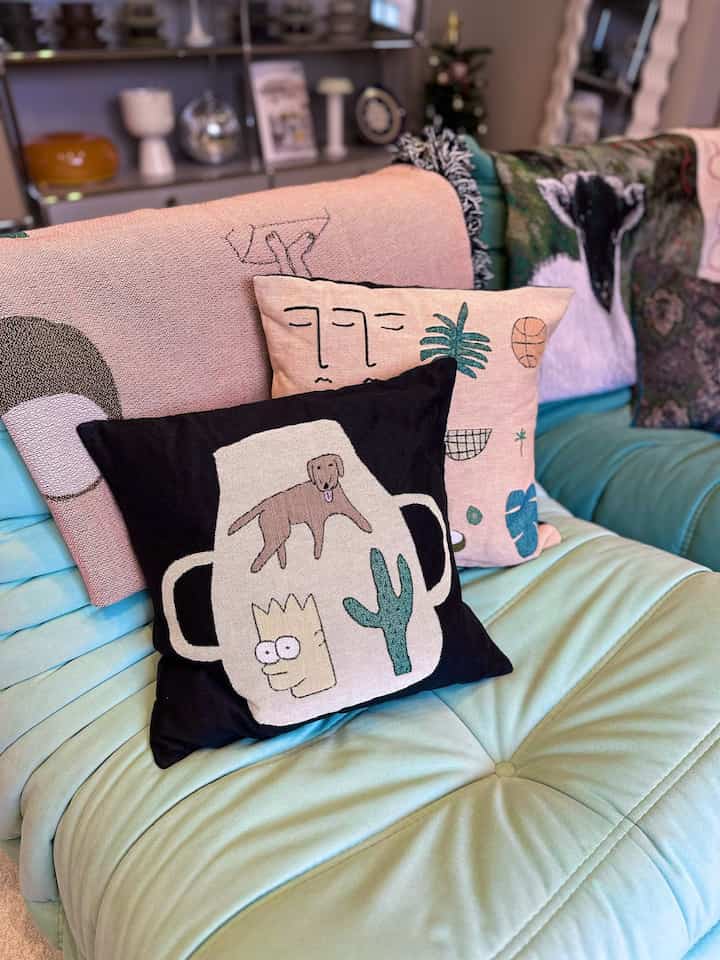 柔らかなアクアトーンのソファに多彩な柄のアートクッションとブランケットが置かれた温かみのあるリビング