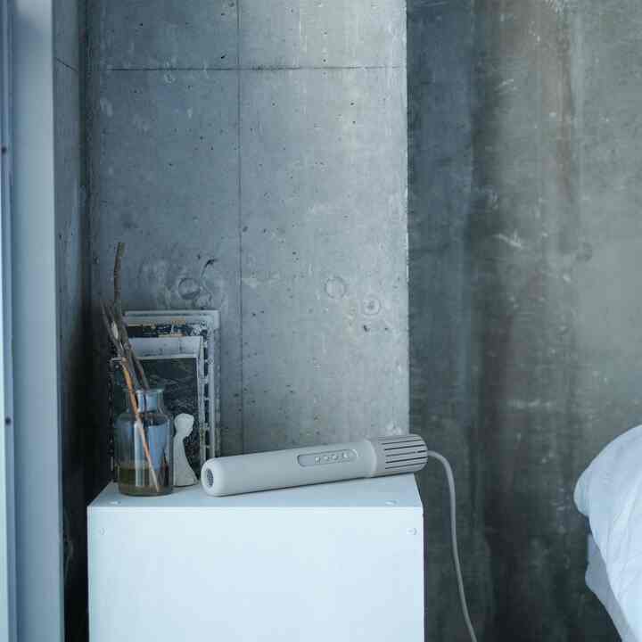 グレーのトーンが印象的な寝室、シンプルなデザインの白いスツールの上にcadoブランドのグレー色のふとん乾燥機とリードディフューザーが置かれた落ち着いた空間
