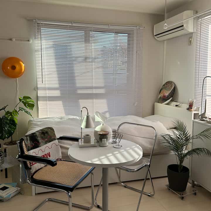 A warm small studio bedroom in white and natural tones featuring window blinds, folding chairs, round table, and a fake plant