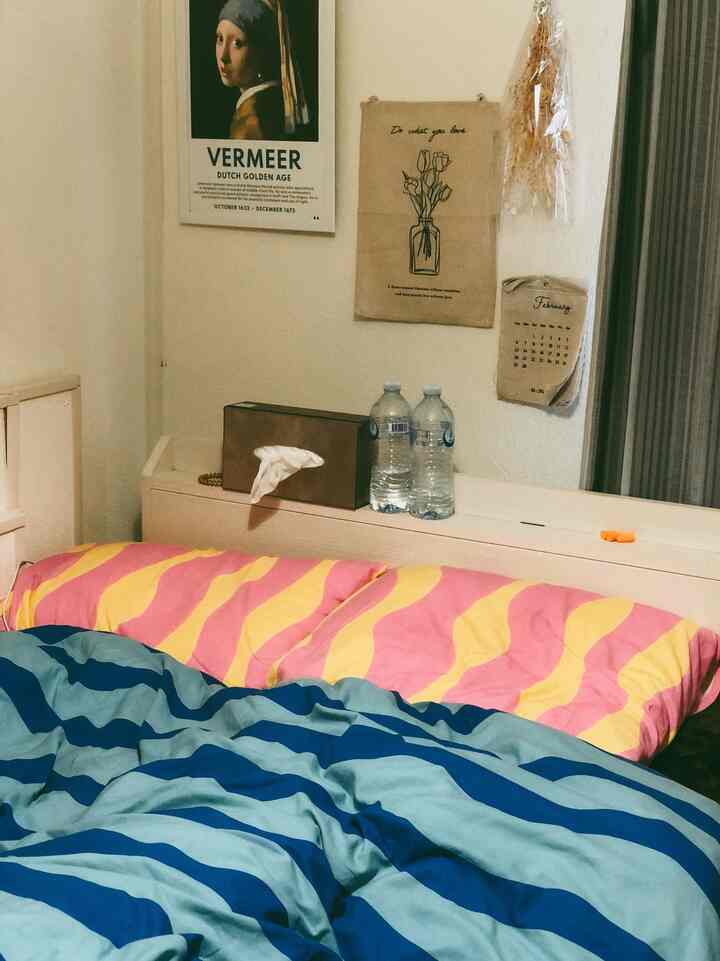 ピンクと黄色のストライプのベッドカバーが置かれたワンルームの寝室、自然な壁飾りとシンプルな家具が調和した雰囲気のあるインテリア