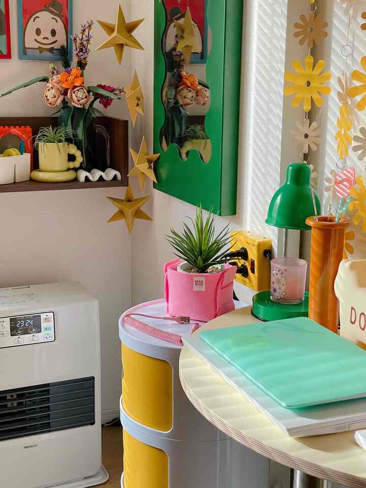 明るくカラフルなワンルーム、ピンクのフェルト製プランターカバーに植物が置かれ、黄色の収納と緑色のテーブルランプが調和したレトロな雰囲気のインテリア