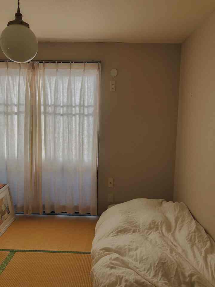ベージュの壁とアイボリーのカーテンがある6畳の狭い寝室、畳の床と白い掛け布団が配置されたシンプルで落ち着いた空間