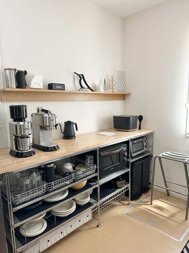 明るいナチュラルトーンのキッチン、ステンレス製ユニット棚とコーヒーマシンが配置されたミニマルで清潔感のあるインテリア