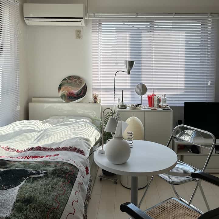 Bright white and natural toned bedroom featuring a round table, transparent stools, and light-filled blinds creating a cozy atmosphere