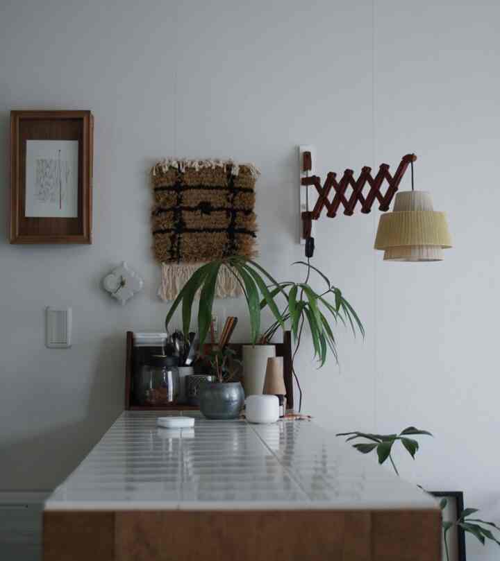 白いタイルのカウンターと木目調の額縁が配置されたナチュラルなキッチン空間、植物と壁照明が調和したカフェ風インテリア