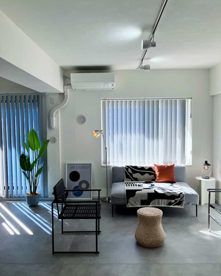 明るいナチュラルトーンのリビング、グレーのソファと黒いラタンチェア、床にラタンの円形スツールとフロアランプが配置されたシンプルモダンなインテリア