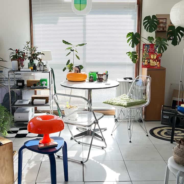 Mid-century modern studio living space featuring white blinds, a white round dining table, transparent chairs, and an orange table lamp