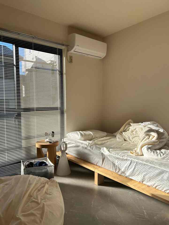 White and natural wood toned bedroom featuring bed, wooden stool, folding laundry basket, and blinds creating a sense of openness