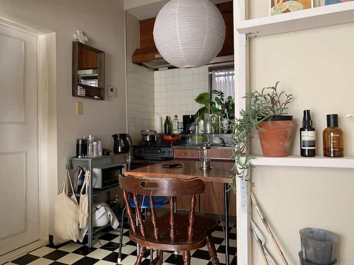 Small kitchen space in white and brown tones, featuring wooden dining table and chairs, shelves with plants, vintage style