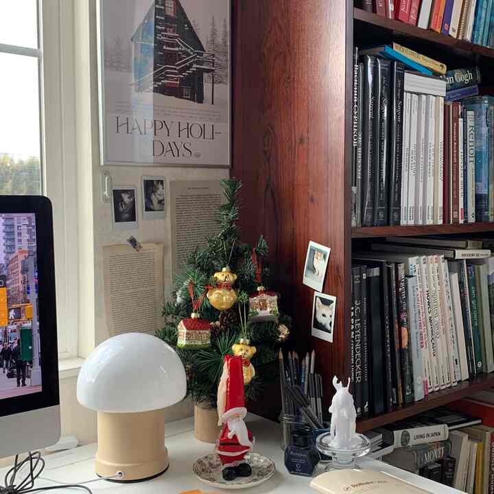 A home office space with white and dark brown tones, featuring a poster, Christmas decorations on desk, and a bookshelf filled with books