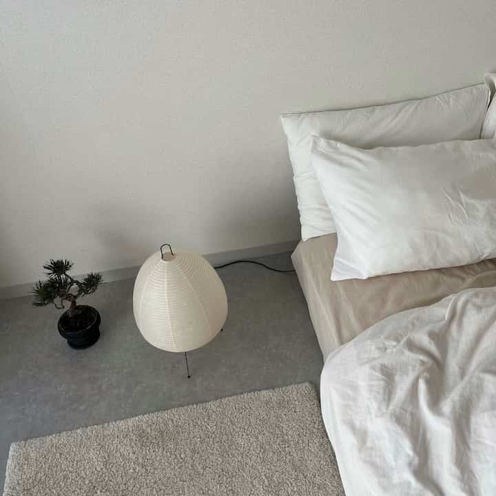 白とグレーのトーンのワンルーム寝室、布団と枕、アカリのフロアランプ、小さな盆栽が配置されたミニマルで自然な雰囲気のインテリア