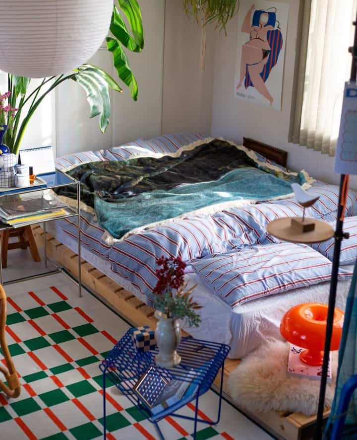 青と赤のストライプ寝具が置かれた寝室、ナチュラルな木製家具と緑の植物が調和した雰囲気のあるインテリア