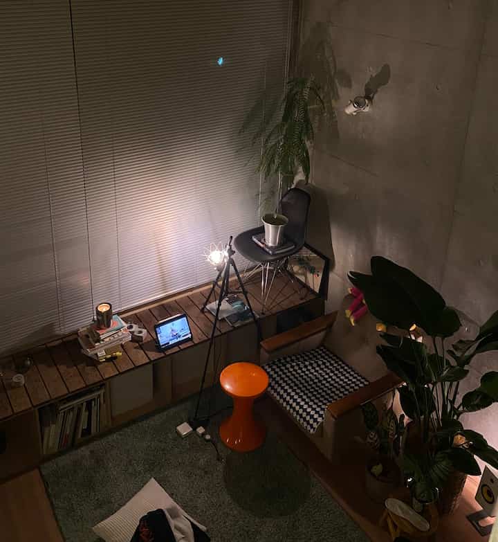 Dark-toned studio apartment featuring wooden shelves, an orange stool, and plants creating a cozy home cafe atmosphere