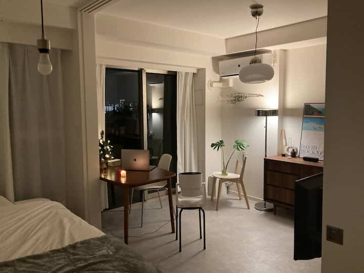 A compact studio apartment in white and brown tones featuring wooden dining table and chairs, a floor lamp, and natural modern decor