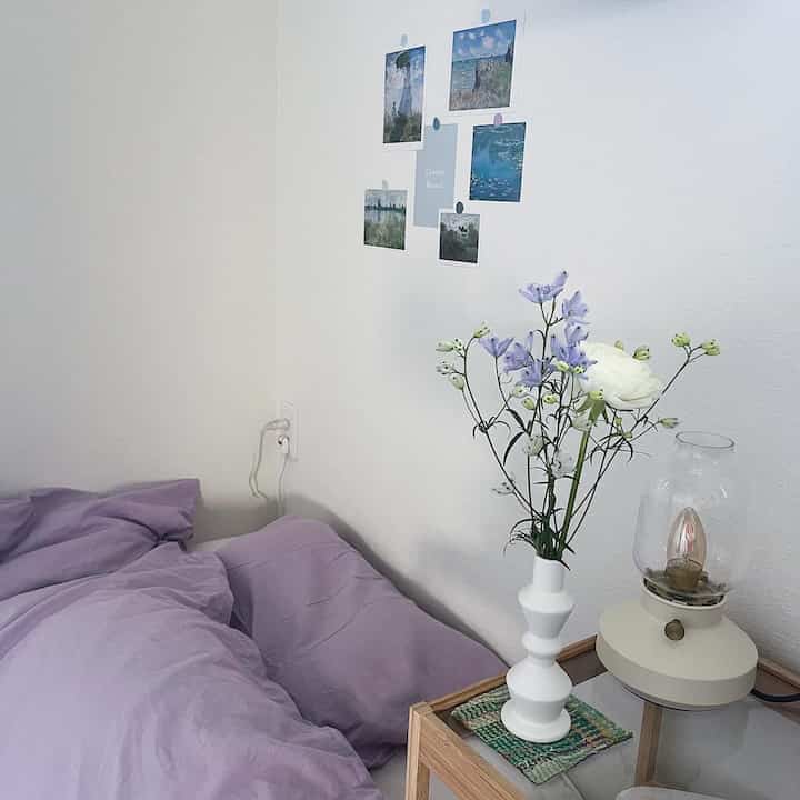 白い壁とラベンダー色の寝具があるシンプルな寝室、ナイトテーブルの上に花瓶とテーブルランプが配置された印象的な空間