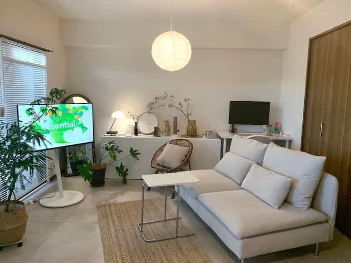 Bright white natural-toned 1LDK living room featuring sofa, pendant light, desk, and green plants creating a modern atmosphere