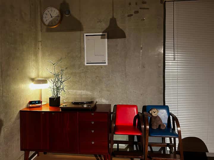 チャコール色のコンクリート壁が背景の北欧風リビング、レコードプレーヤーやテーブルランプ、木製サイドボード、赤と青の椅子、ぬいぐるみの猿が配置された印象的な空間