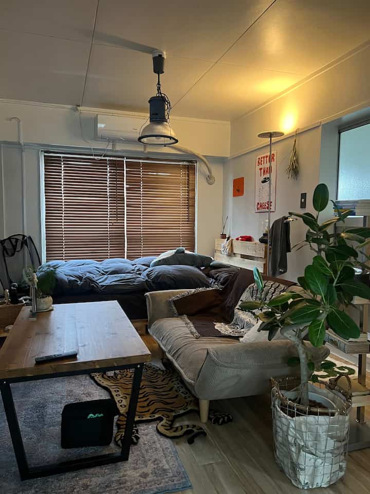 Natural wood tone living room featuring a sofa, low wooden table, bed near window with wooden blinds, and floor lamp with warm ambiance