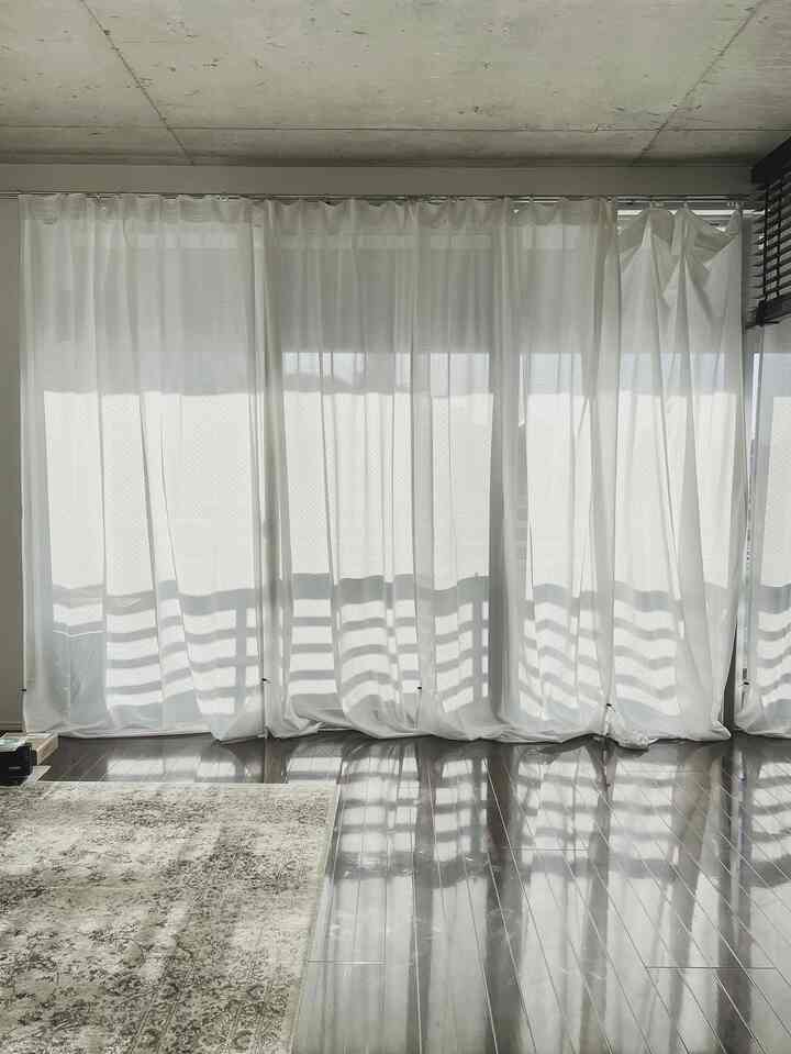 白いカーテンとグレーのラグが配置された木目調の床のナチュラルなリビング、バルコニーを臨む空間