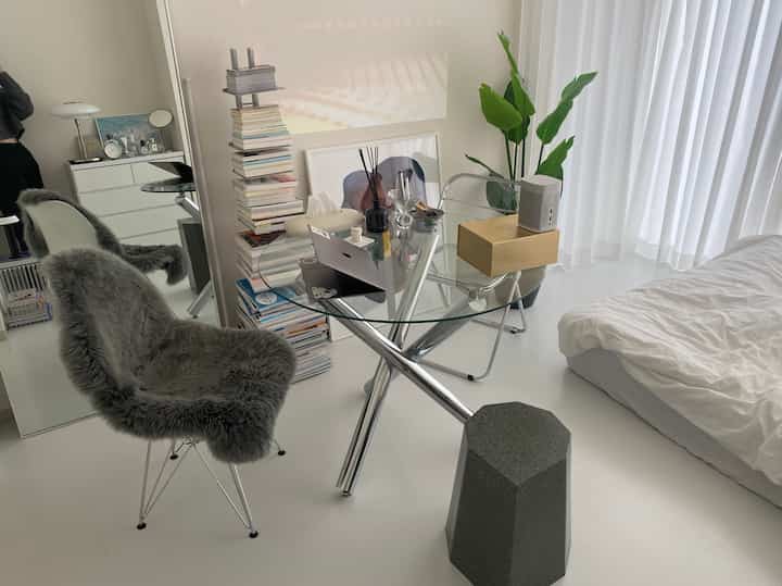 White-toned studio apartment featuring a glass table desk, bookshelf, and plant with a minimal and natural atmosphere