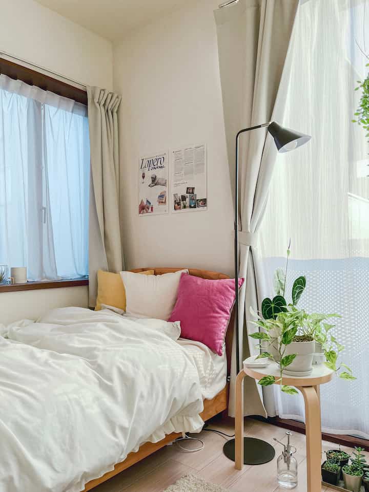 白い寝具と黄色・ピンクのクッションが置かれた居心地の良い一人暮らしの寝室、窓辺に植物が配置された自然な雰囲気のインテリア