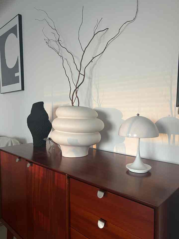 柔らかな白い壁と茶色の木製チェストの上に個性的な形の白い花瓶とテーブルランプが置かれたミニマルなリビングインテリア