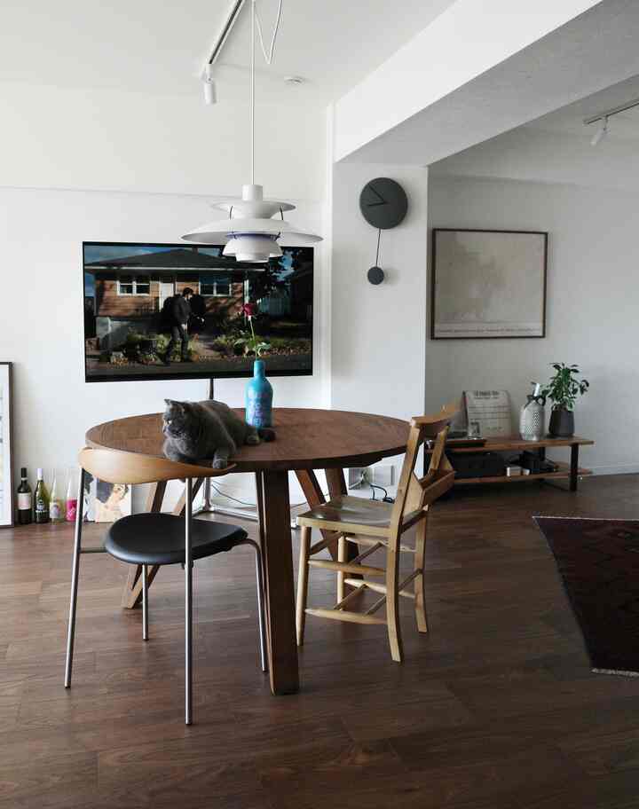 A white-walled living room with wood-toned furniture, featuring a round dining table with a cat sitting on it and a minimalistic atmosphere