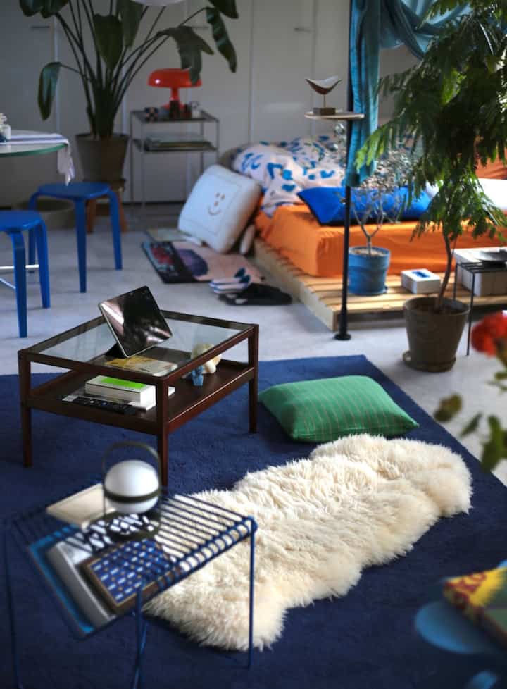 A natural and Mid-Century Modern living room featuring a blue rug, wooden bed frame, blue stools, and indoor plants creating a cozy atmosphere