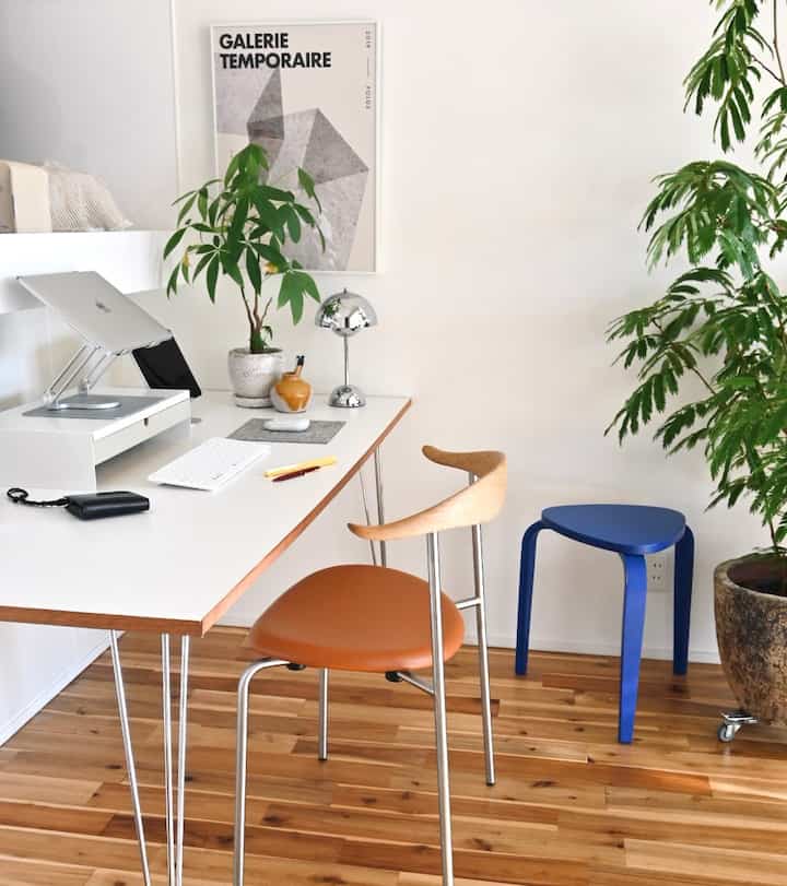 A white-walled workspace with wood-tone flooring featuring a natural wood-edged desk, stools, and plants creating a minimal and natural atmosphere