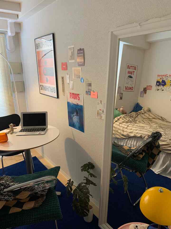 白い壁と青いカーペットが調和する小さなワンルーム、 大きな鏡とデスク、かわいらしいインテリア要素が配置されたシンプルな空間