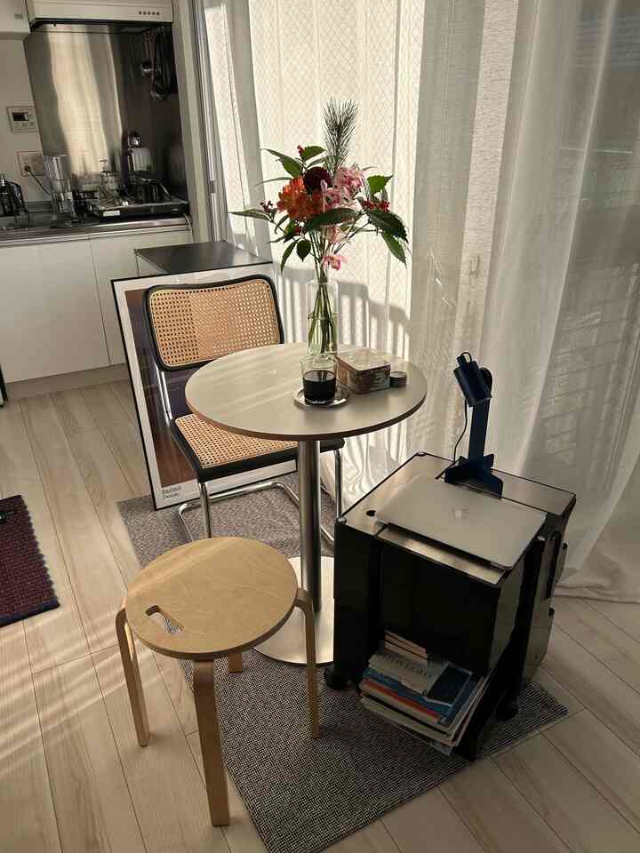 明るいホワイトトーンのコンパクトなリビング兼キッチンスペース、丸いサイドテーブルとラタンチェア、木製スツール、花の装飾が配置された居心地の良いおうちカフェ風インテリア