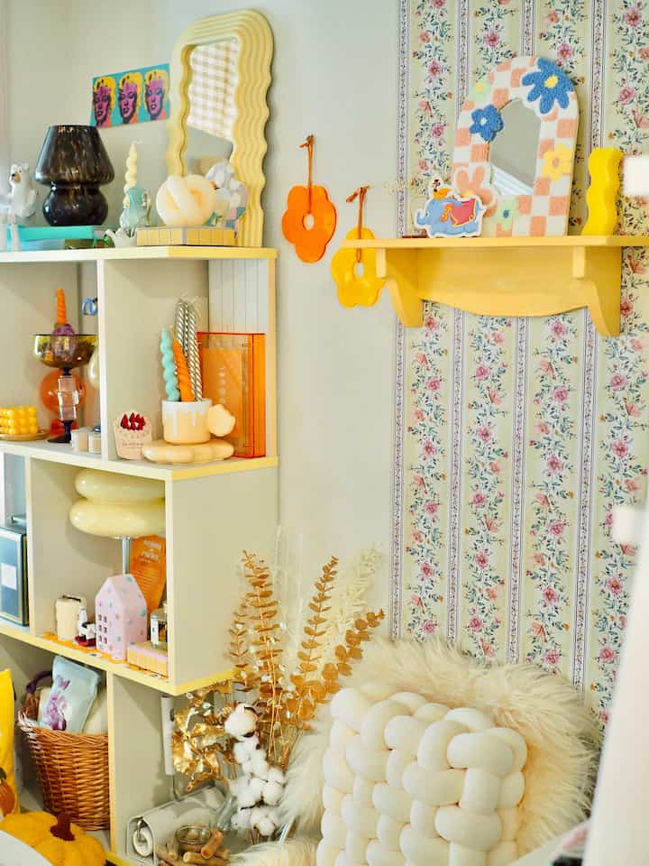 A cozy living room corner featuring a yellow wall shelf, retro floral wallpaper, white braided cushion, and assorted decor items
