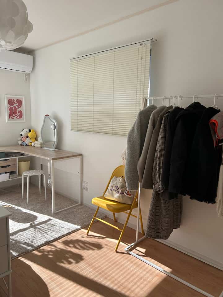 白とベージュのトーンの一人暮らしの部屋、シンプルなデスクとハンガーラック、黄色い折りたたみ椅子が配置された落ち着いた雰囲気のインテリア