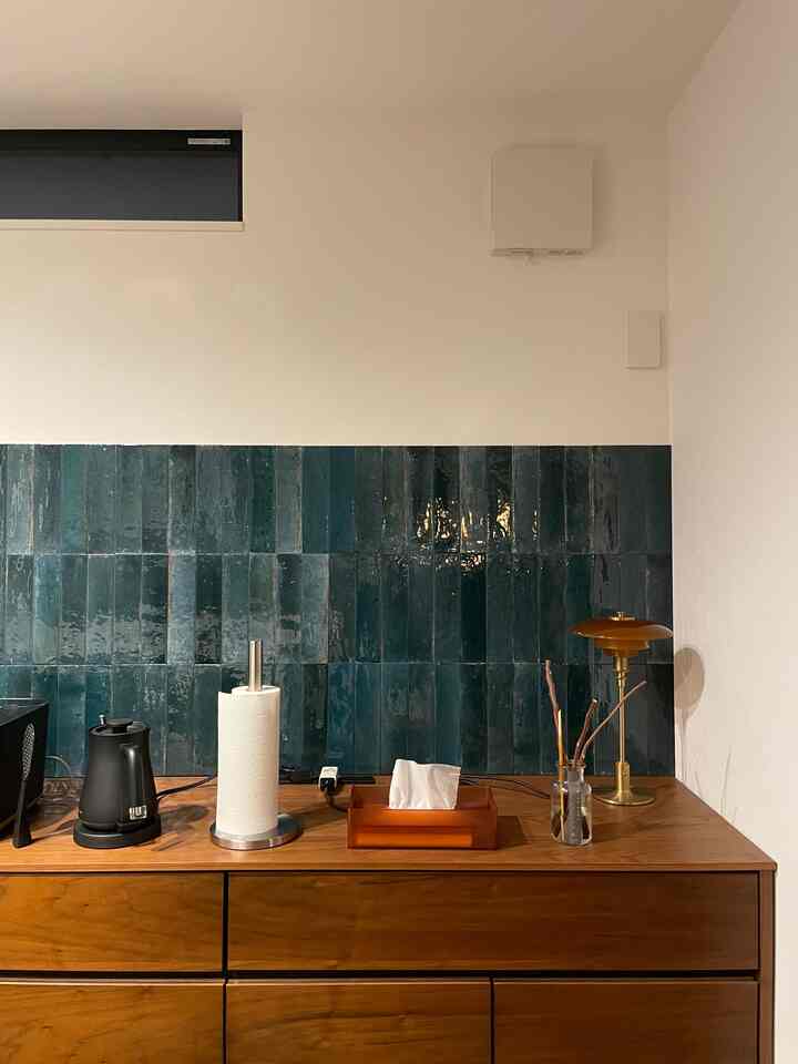 白い壁と濃い青緑色のキッチンタイル、木目調のキッチンスペースにバルミューダの電気ケトルと琥珀色のテーブルランプが配置されたモダンなインテリア