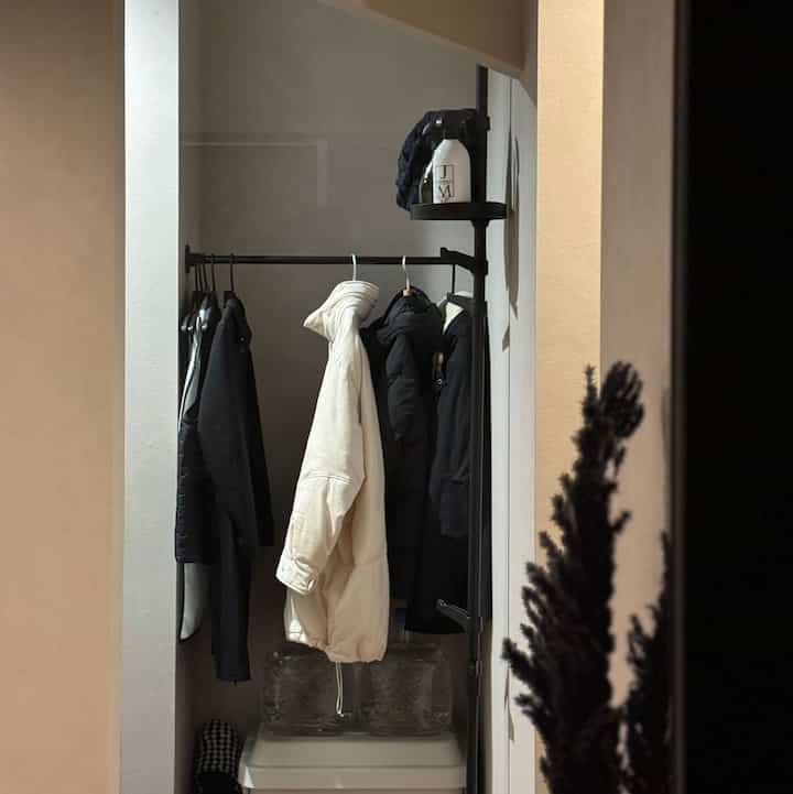 ナチュラルなベージュ色の玄関の一角に、黒い鉄製ハンガーに複数のコートが掛けられた実用的な収納スペース
