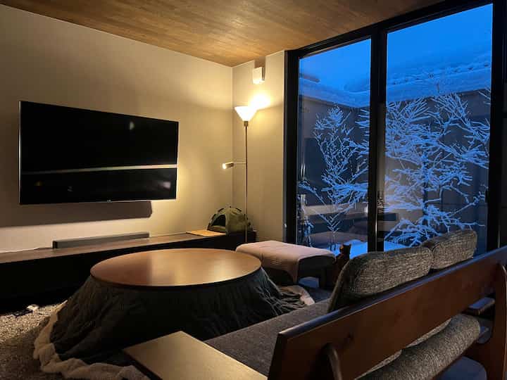 Warm wood-toned living room with soft lighting, featuring a large window showcasing snowy trees outside and a cat inside