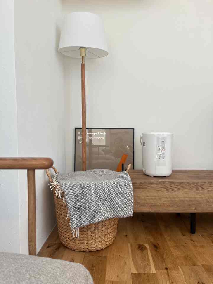 ナチュラルなブラウンの木目調リビングコーナー、白いフロアランプ、テレビ台の上の白い加湿器、ライトブラウンの床、グレー柄のスローが置かれたバスケットのある落ち着いた空間