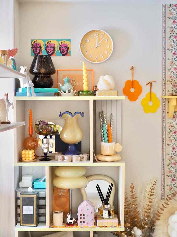 明るい黄色の壁掛け時計とカラフルなキャンドル、花瓶が並ぶ白い棚のある雰囲気の良いリビング空間