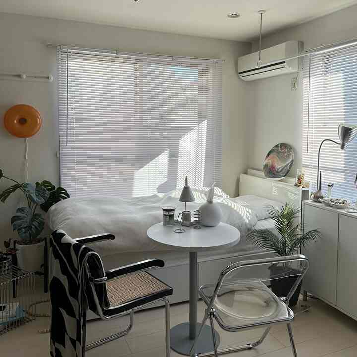 明るい白を基調としたワンルームの寝室、透明な椅子とナチュラルな植物が配置されたモダンなインテリア