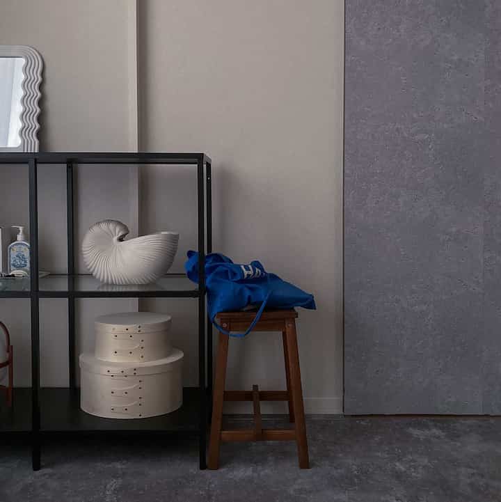 グレーのモノトーンと木目調が調和したシンプルなワンルーム、DIYのモルタル調ドアとミニマルな棚が配置された雰囲気のあるインテリア