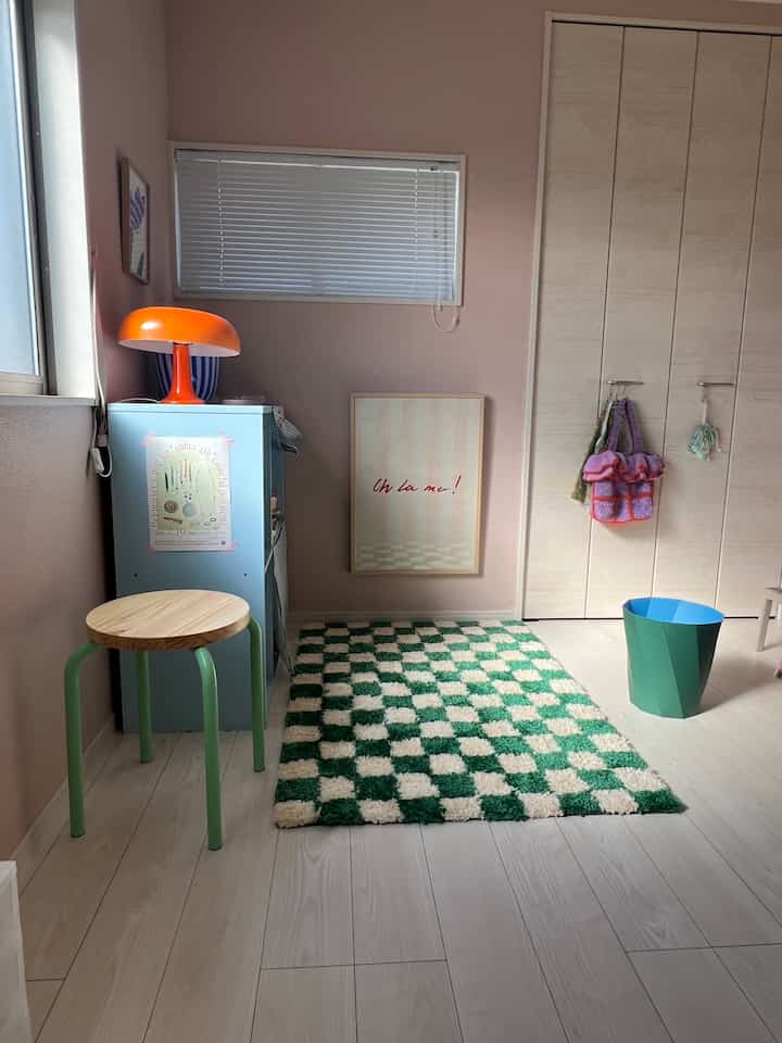 A small rest area with pink-toned walls and green checkered rug, featuring a stool and orange table lamp creating a cozy atmosphere
