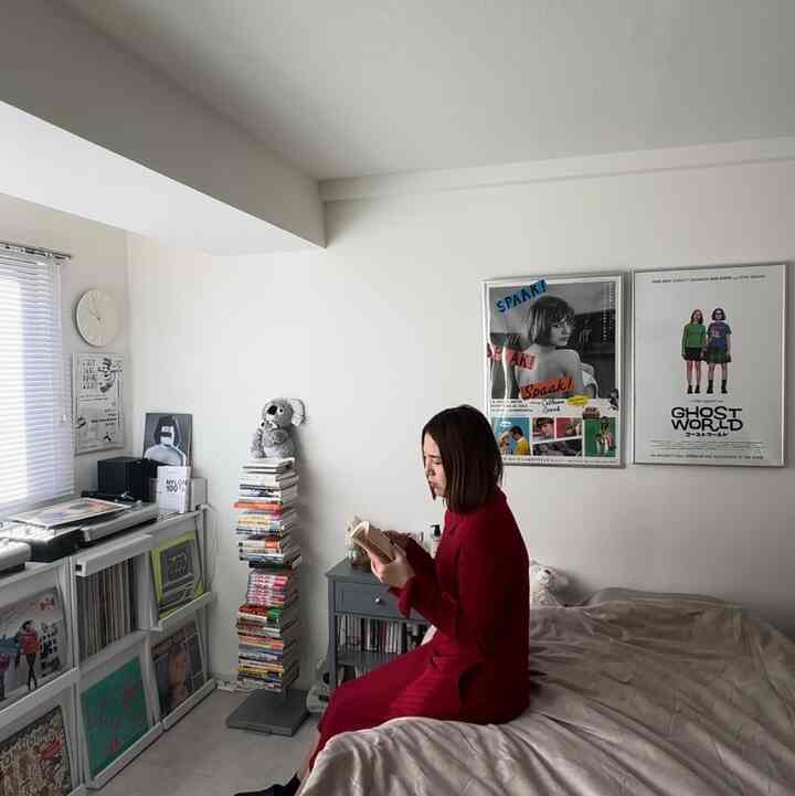 White-toned bedroom featuring bookshelf and book tower, with a woman in red reading, creating a calm atmosphere